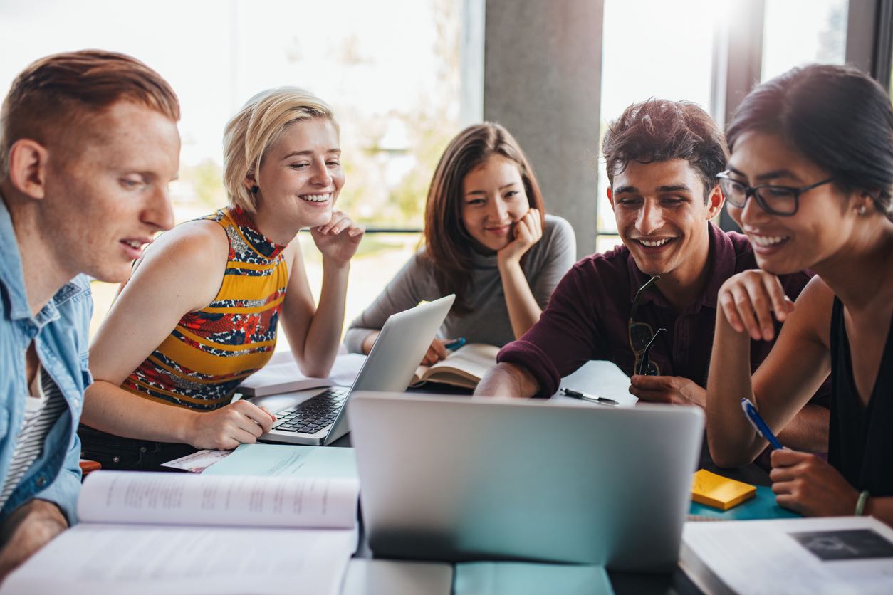 Positive excited multi-ethnic students in casual clothing lying on floor in campus library and laughing while watching curious video on laptop